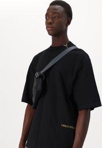 Sort oversized T-shirt med korte ærmer, der har et gult logo detalje ved halsen og en lille, fastgjort sort pouch med en grå rem.