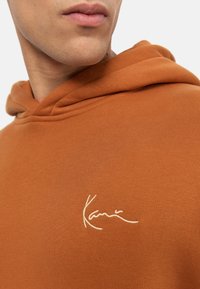 Felpa marrone realizzata in un tessuto texturizzato, con cappuccio regolabile e logo bianco ricamato sul petto.