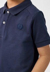 Marineblauw poloschirt met korte mouwen, gemaakt van gestructureerde stof. Heeft een ribbelkraag en een ronde marineblauwe logopatch op de linkerkant van de borst.
