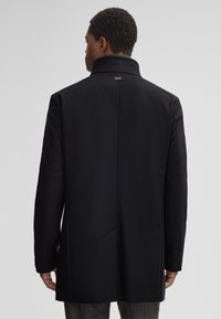 Uomo visto da dietro che indossa un cappotto nero con colletto alzato e pantaloni grigi, su uno sfondo chiaro e semplice.
