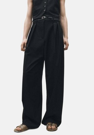 Black wide-leg trousers with a high waist, featuring pleats, a belt, and a relaxed fit. Paired with patterned sandals.