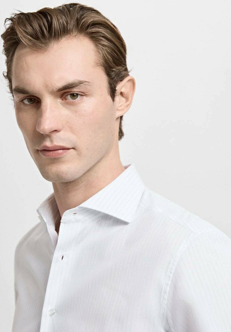 Chemise blanche à rayures verticales subtiles, avec un col structuré et des détails de boutons. Le tissu semble net et lisse.