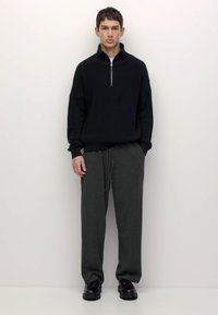 Fiatal férfi áll egy fehér falnak támaszkodva, fekete félcipzáros pulóverben, szürke, kötéllel húzott nadrágban és fekete cipőben.