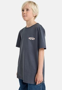 T-shirt en coton gris foncé à manches courtes, présentant un petit patch logo rouge et blanc sur la poitrine gauche. Design minimaliste, coupe décontractée.