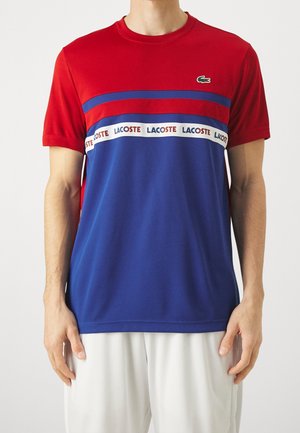 Mann trägt rotes und blaues kurzärmeliges T-Shirt mit Lacoste-Logo-Band quer über der Brust und grünem Krokodil-Emblem oben links.