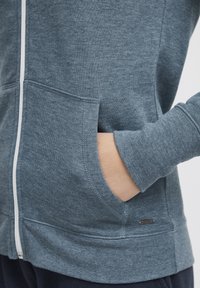 Aizdaru džemperis tumši pelēkzilā audumā, ar sānu kabatām, ribotiem piedurkņu galiem un nelielu logotipa akcentu apakšējā pusē. Mīksta tekstūra.