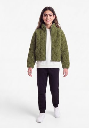 Fille souriante aux longs cheveux portant une veste zippée verte texturée, une chemise blanche, un pantalon noir et des baskets blanches, debout sur fond blanc.