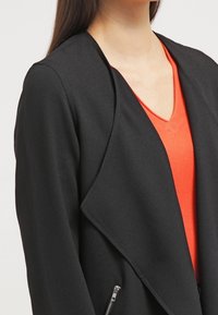 Giacca nera con fronte drappeggiato, texture liscia e dettaglio con zip argentata di lato, indossata sopra una maglietta arancione con scollatura a V.