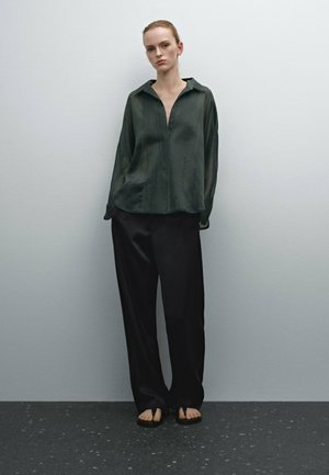 Dark green, textured, long-sleeve blouse with a collar, paired with loose-fitting, black trousers. Flat sandals complete the outfit.
