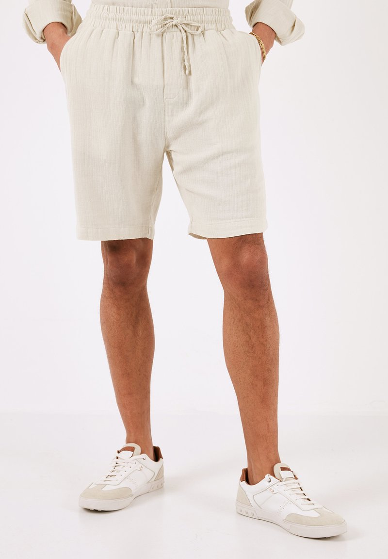 Shorts in beige chiaro con texture, dotati di una fascia elastica in vita e cordoncino, abbinati a scarpe da ginnastica bianche con dettagli grigi chiari.