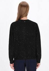 Pulover negru cu un design texturat, cu accente argintii dispersate, cu manșete și tiv ondulate și un gât rotund. Vedere din spate.