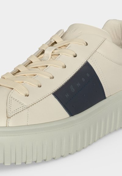Gros plan d'une basket crème avec une semelle épaisse, des lacets crème et un patch bleu foncé affichant un texte pâle sur le côté.