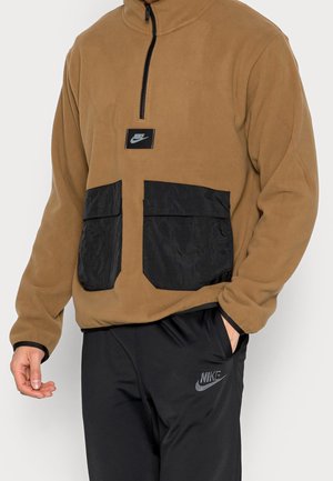 Bruine fleece pullover met een ritskraag, zwarte nylon voorzakken en een klein Nike-logo patch. Gecombineerd met zwarte sportbroek.