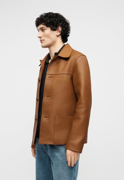 Uomo con capelli ricci scuri che indossa una giacca di pelle marrone sopra una maglietta nera e jeans blu, in piedi di lato contro uno sfondo neutro.