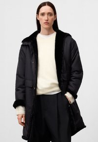 Kvinna klädd i en lång svart kappa med pälsdetaljer över en vit tröja och svarta byxor, stående framför en enfärgad ljus bakgrund.