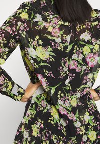 Kukkamekko, jossa on musta läpikuultava kangas, jossa on vaaleanpunaisia, keltaisia ja valkoisia kukkia, pitkät hihat, röyhelöhelma ja vartalonmyötäinen siluetti.