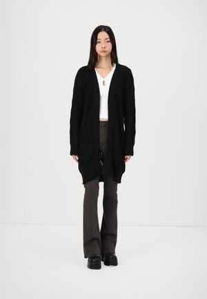 Black knitted cardigan with a relaxed fit, featuring a textured pattern and open front. Paired with a white top and dark pants.