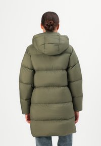 Vrouw met een donkere gevlochten knot, gekleed in een olijfgroen gewatteerd pufferjack met een capuchon, staande tegen een effen witte achtergrond.