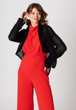 Kobieta pozująca w czerwonym, bezrękawnym kombinezonie i czarnym ażurowym kardiganie, uśmiechająca się, z jedną dłonią przy uchu, a drugą na biodrze.
