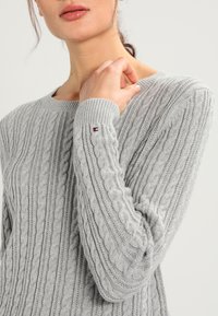 Pull gris en maille torsadée avec un col rond, des poignets côtelés et un petit logo en accent. Le tissu semble doux avec un motif texturé.