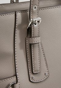 Gros plan d'une sangle de sac en cuir texturé gris avec une boucle métallique argentée et une couture détaillée sur le bord du sac.