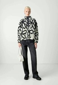 Schwarz-weißer Fleece-Pullover mit floralem Muster und hohem Kragen, kombiniert mit dunklen Jeans und schwarzen Sneakers, während eine weiße Handtasche gehalten wird.