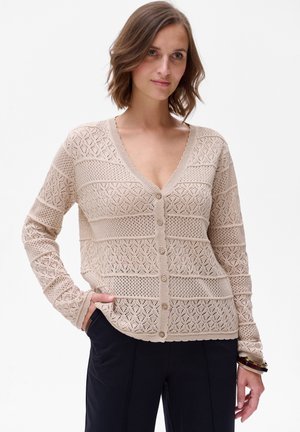 Beige gebreide cardigan met een opengewerkt patroon, diepe V-hals, knopen aan de voorkant en lange mouwen, gecombineerd met donkere broeken.