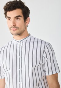 Camicia bianca a maniche corte con colletto, caratterizzata da sottili righe verticali nere, chiusura con bottoni e polsini a finitura piatta.