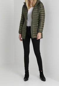 Olivengrøn pufferjakke med hætte, med horisontalt quiltet design og frontlynlås. Parret med sorte tætsiddende bukser og ankelstøvler.