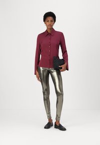 Maroonowa koszula z guzikami, metaliczne złote legginsy, czarna teksturowana kopertówka oraz czarne loafersy. Gładkie materiały, dopasowany krój i elegancki design.