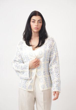 Mujer con cabello negro que lleva un cárdigan de crochet en crema y azul claro sobre una camiseta blanca y pantalones beige, posando frente a un fondo blanco liso.