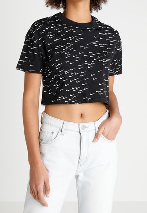 Czarna krótka koszulka z białym wzorem logo Nike, noszona z białymi dżinsami z wysokim stanem, jedna ręka w kieszeni, widoczna środkowa część ciała.