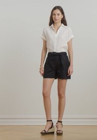 Camisa de linho branca de botão com mangas curtas dobradas, combinada com calções pretos de cintura alta com pregas e um cinto para amarrar. Sandálias espadrille pretas.