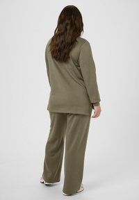 Personne avec de longs cheveux bruns portant un haut à manches longues olive et un pantalon large, se tenant de dos sur un fond blanc.