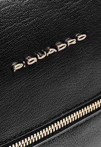 Bolso de cuero negro texturizado con un logo "Piquadro" dorado y una cremallera dorada con detalle de dientes visibles.