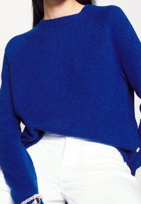 Modrý pletený svetr s kulatým výstřihem, dlouhými rukávy a volným střihem, v kombinaci s bílými kalhotami. Viditelná textura připomínající vlnu.