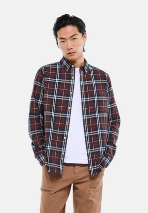Homme portant une chemise à carreaux marron, bleu marine et blanche boutonnée sur un t-shirt blanc avec un pantalon beige, debout les mains dans les poches.