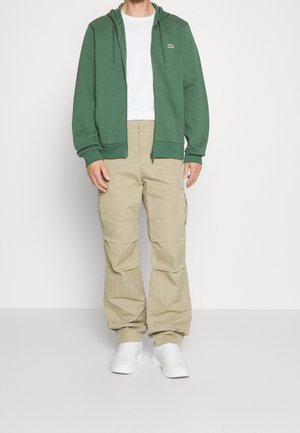 Personne portant un sweat à capuche zippé vert, un t-shirt blanc, un pantalon cargo beige et des baskets blanches, debout devant un fond uni.