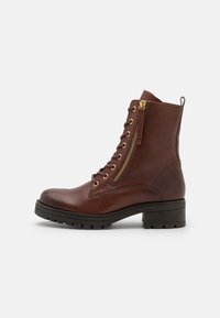 Botas de cuero marrón hasta el tobillo con punta redondeada, cordones en la parte delantera y cremallera lateral. Cuentan con una suela de goma negra resistente y detalles en dorado.