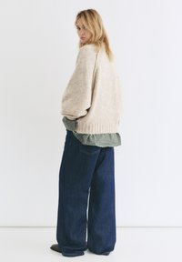 Svetlobežový pletený sveter cez zelenú kockovanú košeľu, v kombinácii so voľnými modrými džínsami, stojac na jednoduchom bielom pozadí.