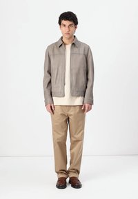 Blusão de couro cinza claro com fecho éclair à frente, t-shirt bege lisa, calças beges e sapatos castanhos. Textura suave e design minimalista.