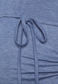 Primer plano de un cordón de tela azul atado en una prenda fruncida con costuras visibles y material de punto texturizado.