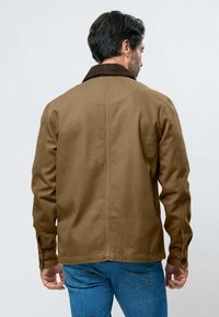 Homme portant une veste marron avec un col foncé et un jean bleu, vu de dos sur un fond clair uni.