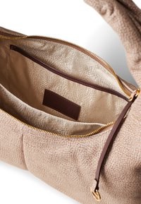 Sac à main beige texturé avec une fermeture éclair dorée, présentant un intérieur à motifs et une étiquette en cuir marron. L'intérieur comprend des poches de compartiment.