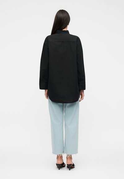 Γυναίκα που στέκεται με την πλάτη, φοράει ένα μακρύ μαύρο πουκάμισο, ανοιχτό μπλε τζιν με ξεφτισμένα τελειώματα και μαύρα πέδιλα με τακούνι και ανοιχτά δάχτυλα, σε λευκό φόντο.