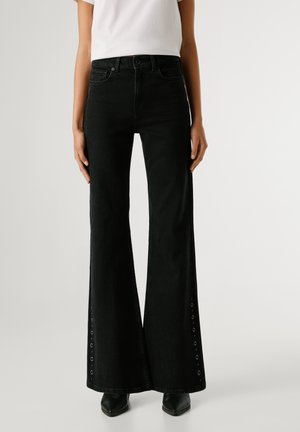 Pantalones vaqueros negros acampanados de mezclilla con cintura alta, que presentan recortes laterales sujetos con ojales de metal y un diseño clásico de cinco bolsillos.