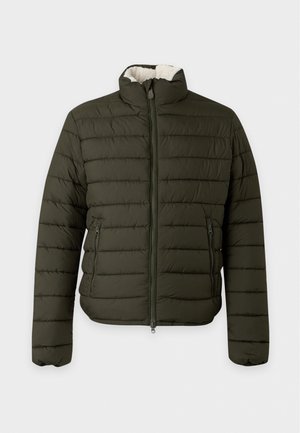 Jeune homme debout, les mains dans les poches, portant une doudoune vert foncé, un t-shirt blanc, un jean bleu large et des baskets blanches sur un fond uni.
