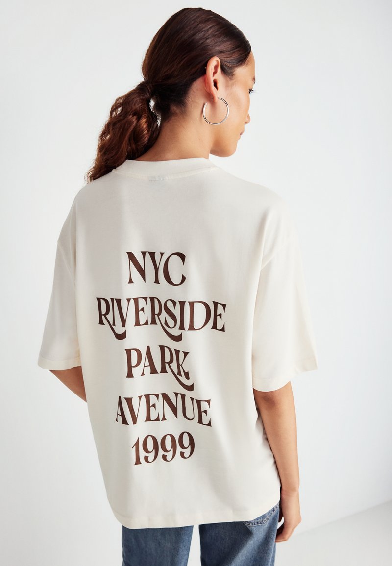 Camiseta de gran tamaño color crema con escote redondo. La parte posterior presenta un texto gráfico marrón: "NYC RIVERSIDE PARK AVENUE 1999". Tejido suave.