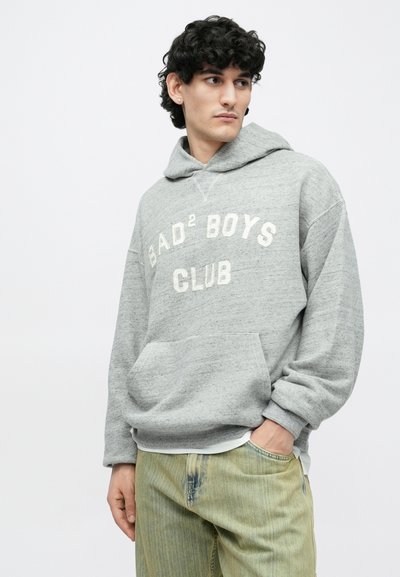 Giovane uomo con capelli ricci scuri che indossa una felpa con cappuccio grigia con la scritta "BAD² BOYS CLUB" e pantaloni beige, in piedi con una mano in tasca.