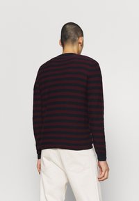 Homme de dos portant une chemise à manches longues rayée noire et rouge foncé et un pantalon blanc cassé sur un fond blanc uni.